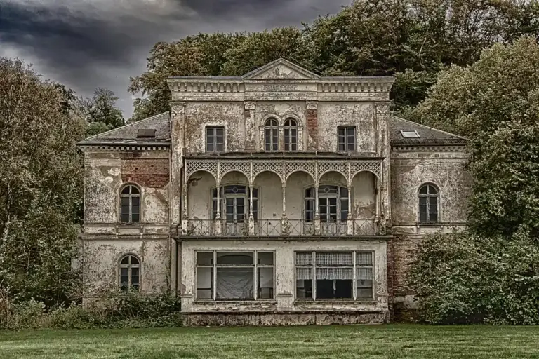 Abandoned two-story mansion with peeling exterior, arched balconies, and overgrown trees and lawn