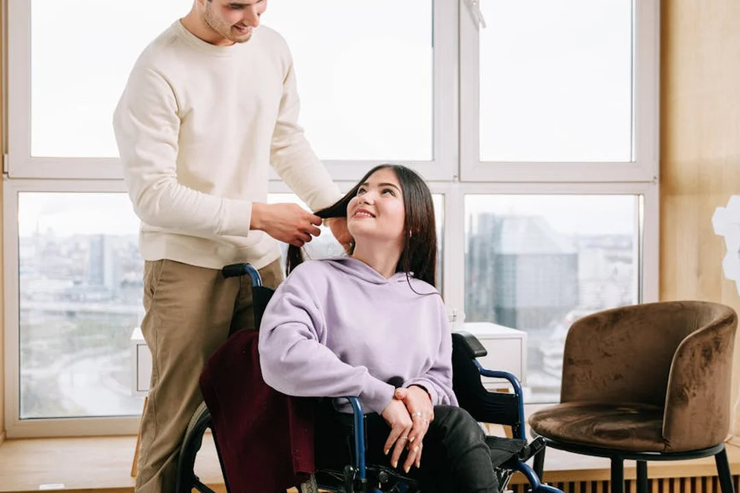A person in a wheelchair in a bright room with large windows, being assisted by a standing companion.