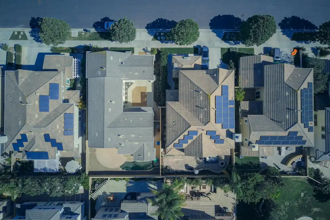 Aerial view of a suburban neighborhood with rows of houses, grey and blue roofs, and surrounding trees.