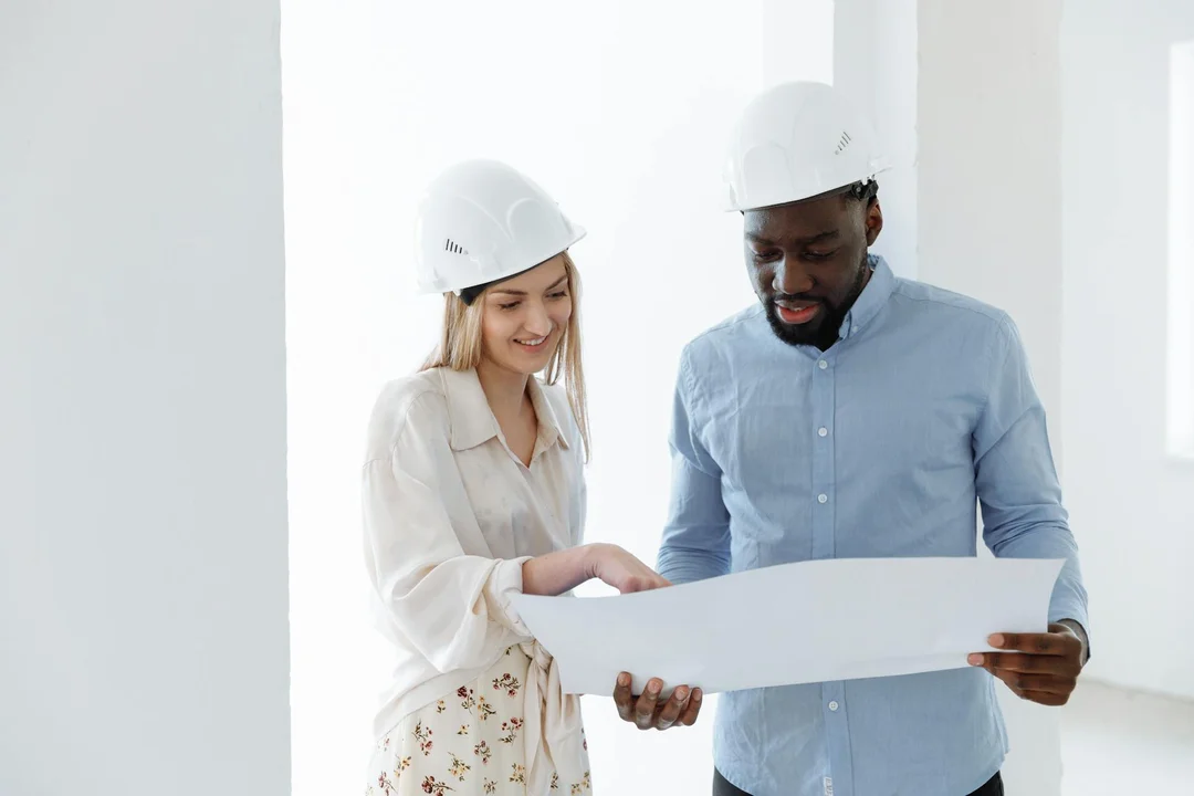 Two professionals wearing hard hats review blueprints in a bright, modern space.