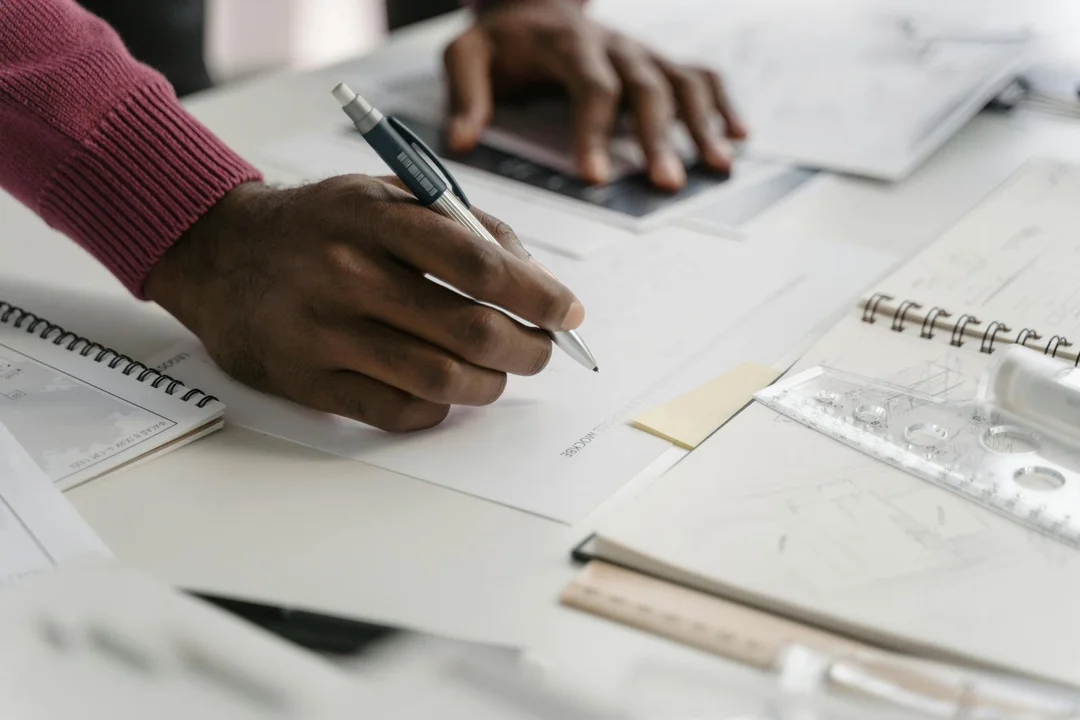 A close-up of hands writing on documents with notebooks and rulers on a desk, preparing evidence for an architectural appeal.