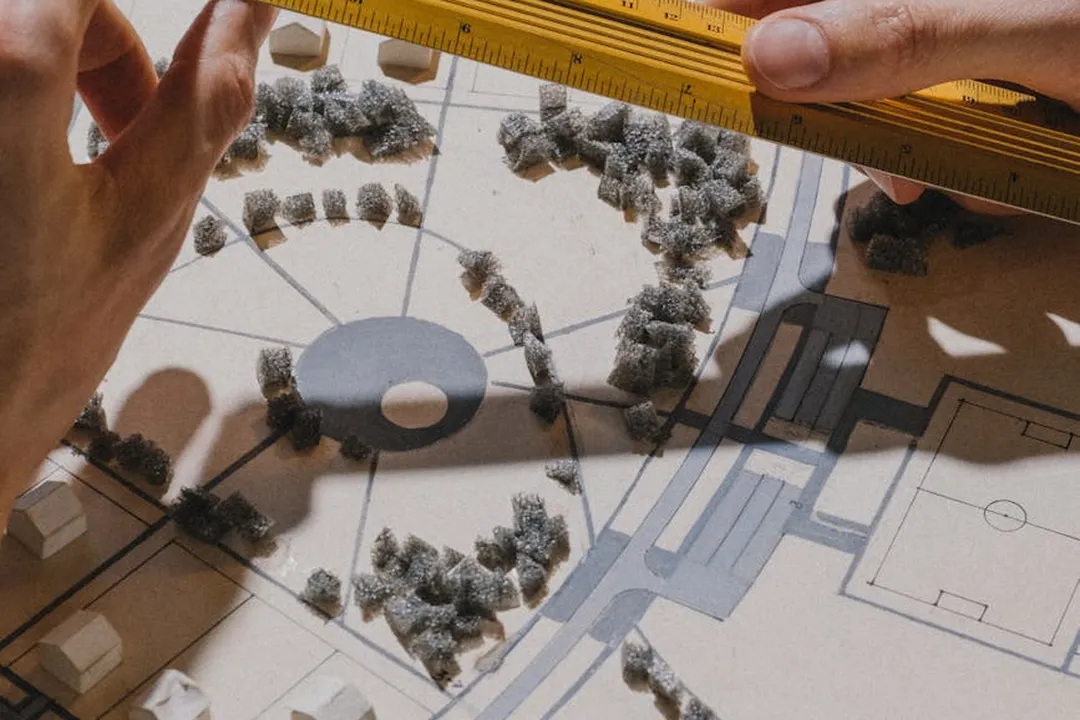 Close-up of hands adjusting a yellow ruler over an architectural plan with miniature trees and buildings.