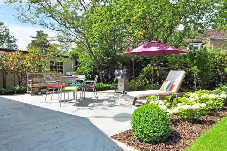 Sunny backyard patio with outdoor seating, a pink umbrella, and manicured greenery.