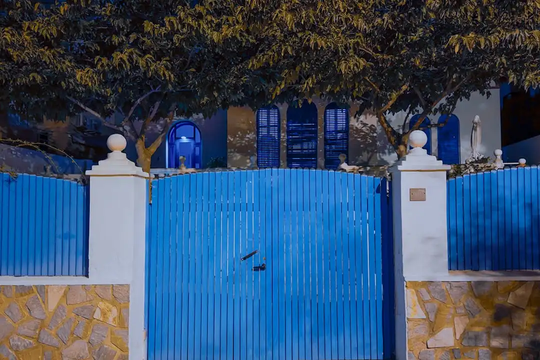 Blue wooden gate of a townhouse community, flanked by white pillars and stone walls with trees overhead.