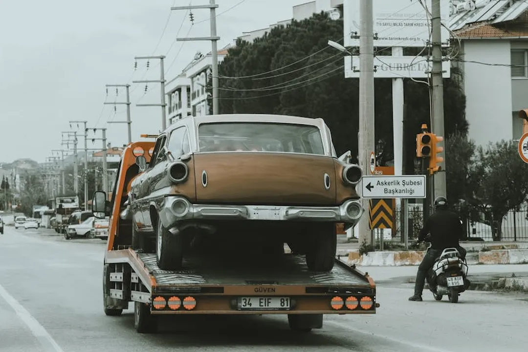 Vintage car being loaded onto a flatbed tow truck on a busy urban street, with signs and motorcycles in the background.