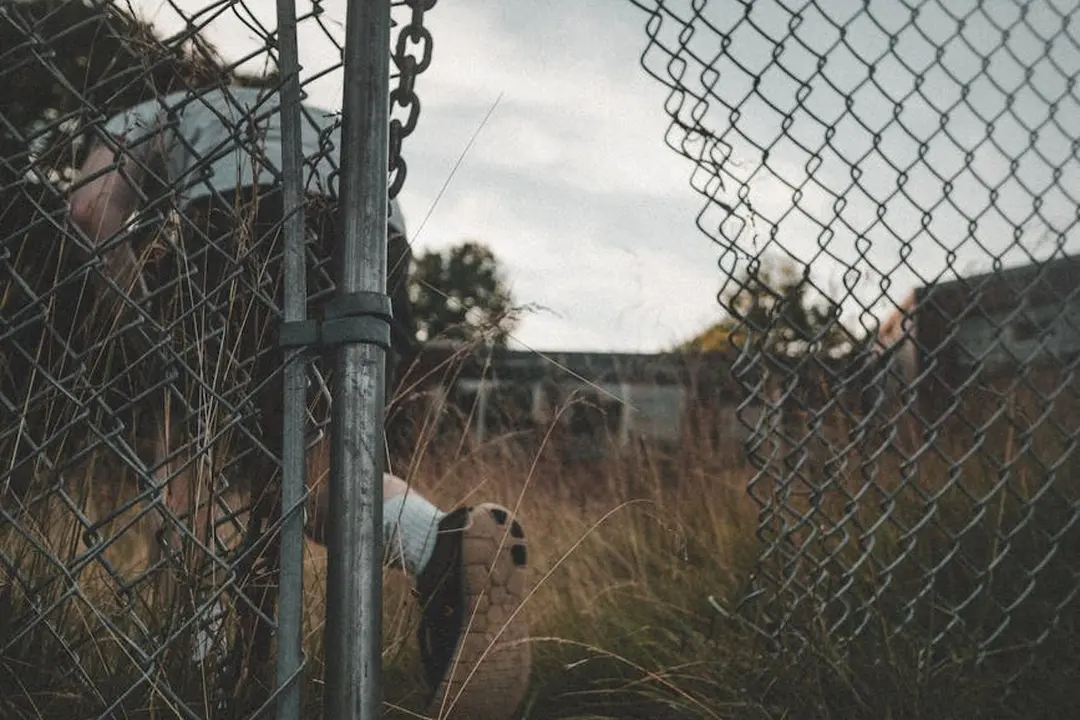 A chain-link fence with a gate and overgrown grass, with someone partially visible behind the fence.