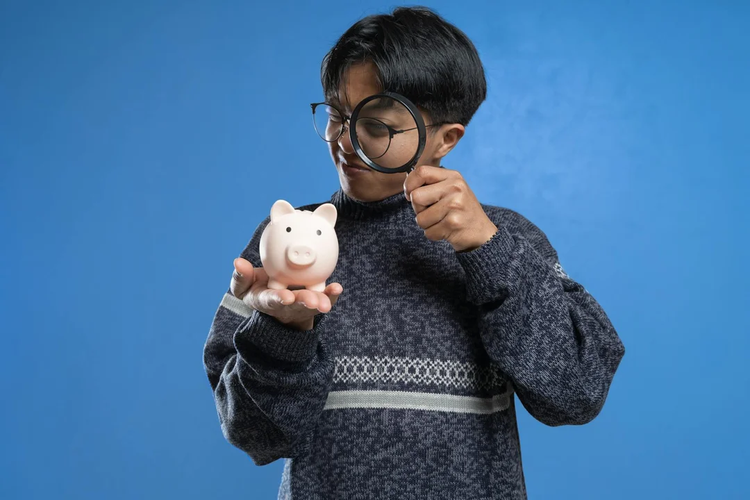 A young child wearing glasses and a dark sweater holds a pink piggy bank, standing against a solid blue background.