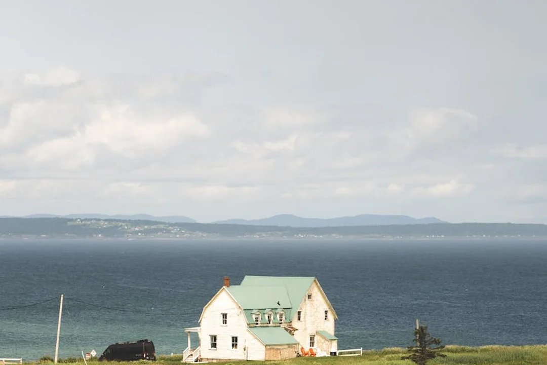 White cottage with a green roof by the sea on a grassy shore under a cloudy sky.