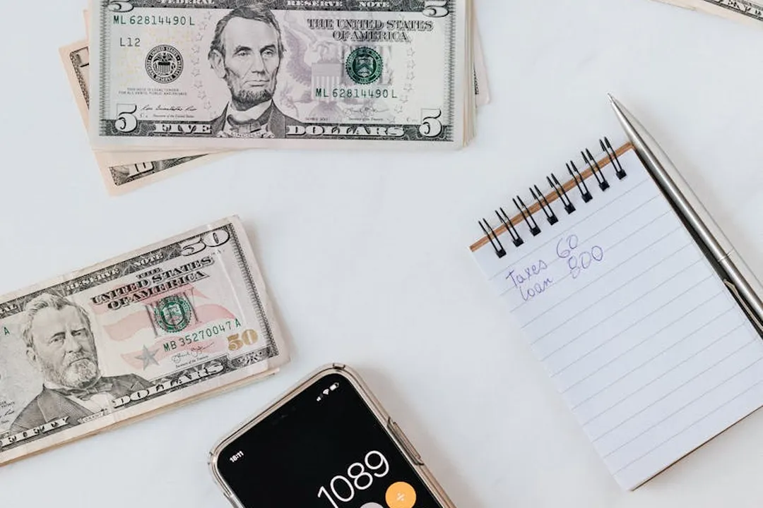 US dollar bills, a small spiral notebook with a handwritten note, and a smartphone on a white surface.