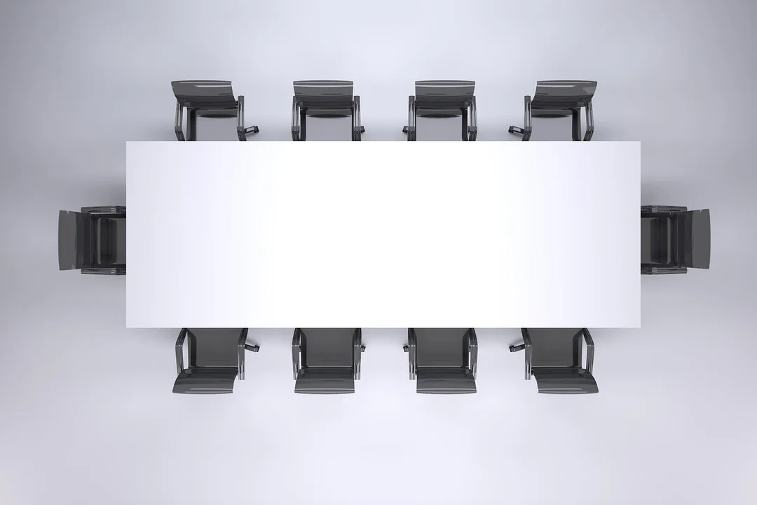 Overhead view of a large rectangular white conference table surrounded by black chairs in a minimalist meeting room.
