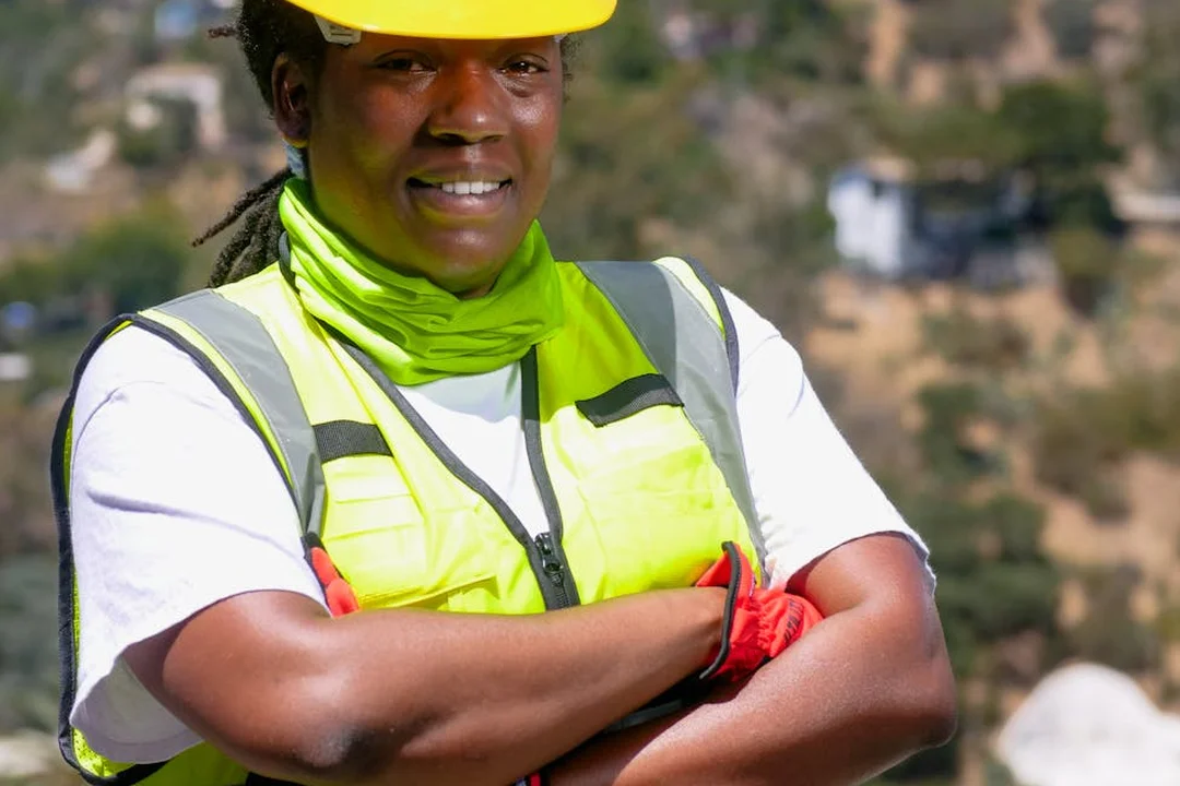 Construction worker wearing a neon safety vest and hard hat standing with arms crossed on an outdoor job site.