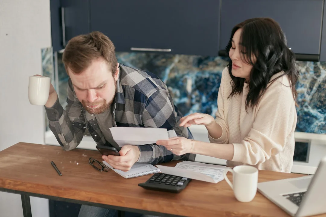 A man and a woman sit at a wooden kitchen table reviewing mortgage documents with a calculator, mugs, and papers, discussing escrow and HOA fees.