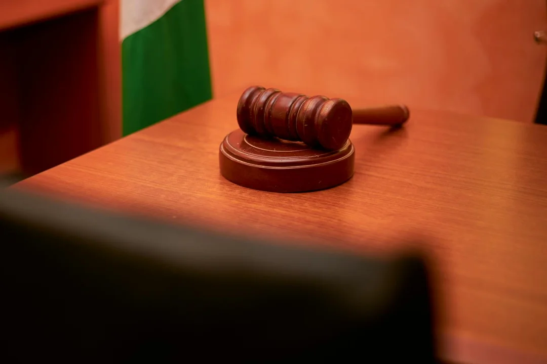 A judge's gavel resting on a wooden courtroom desk, with a blurred foreground and a flag in the background.