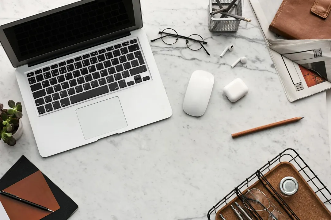 Top-down view of a clean workspace with a laptop, wireless mouse, glasses, notebooks, and office supplies, representing the digital tools used to manage a self-managed HOA.