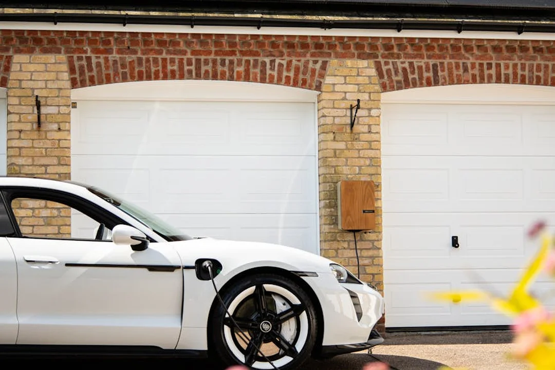 White electric vehicle charging at a wall-mounted charger in front of a brick townhouse with two white garage doors.