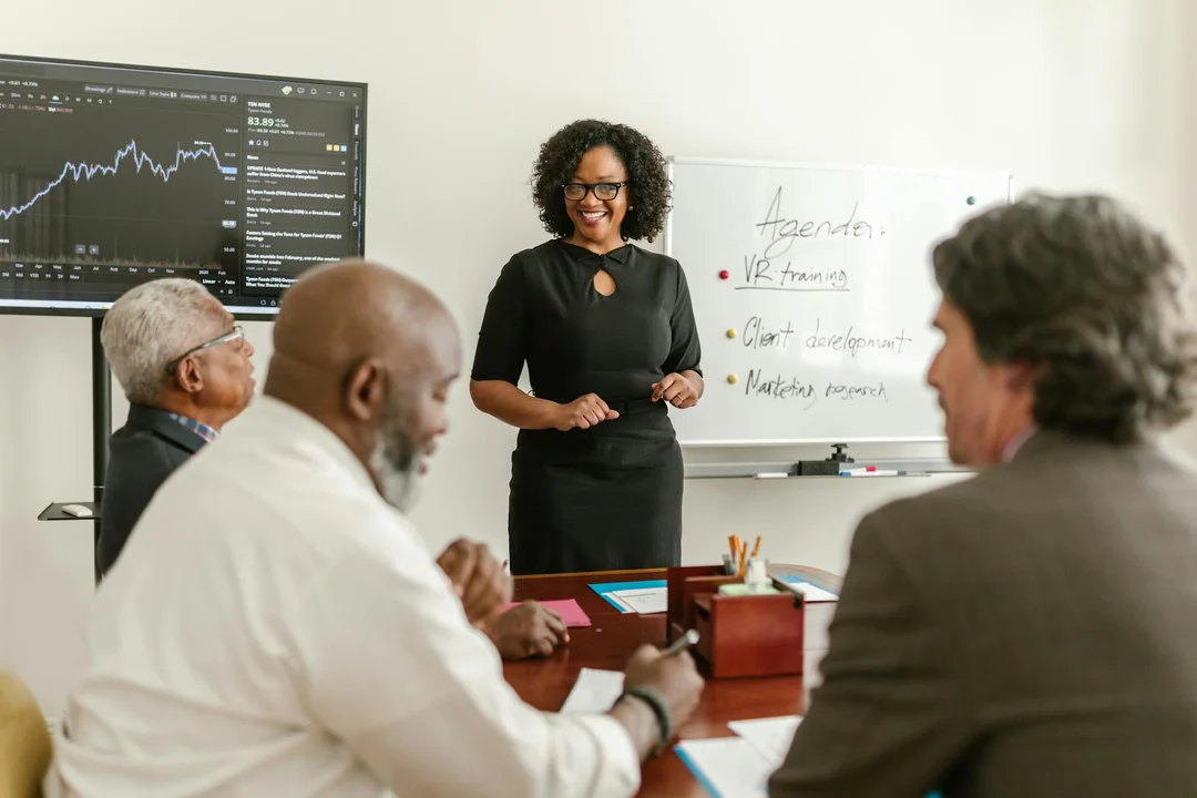 Diverse group of professionals seated around a conference table listening to a presenter in a black dress near a whiteboard; a monitor showing charts is mounted in the background.