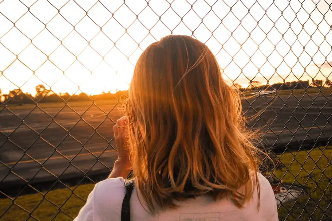 A person with shoulder-length hair stands with their back to the camera, looking through a chain-link fence at a sunset.