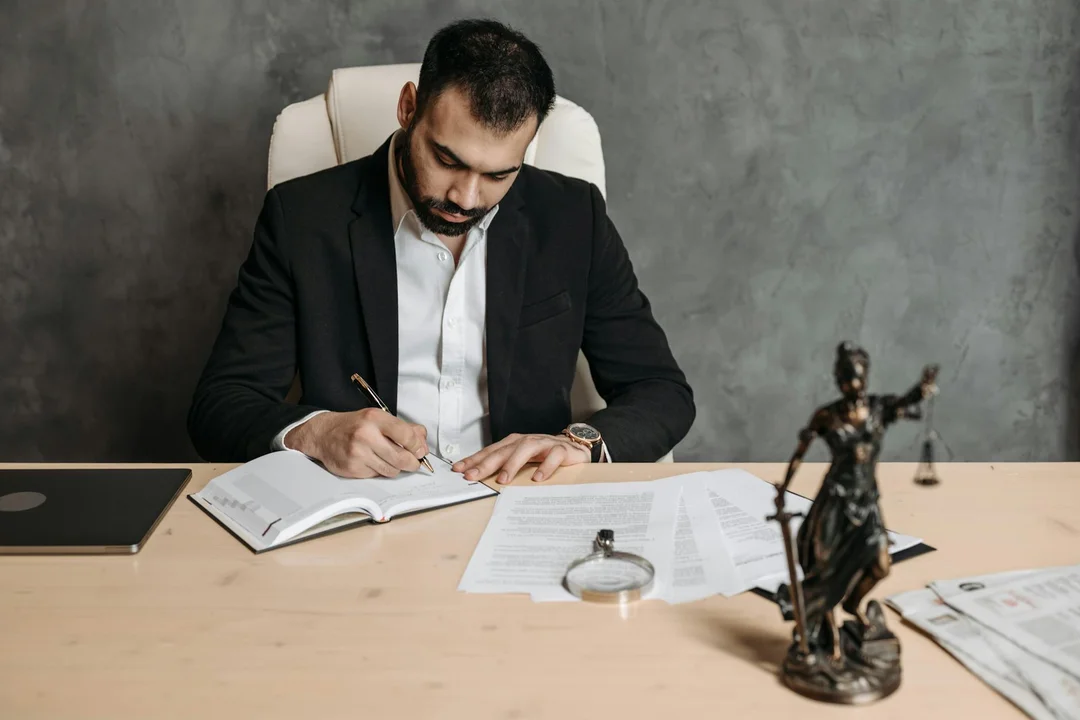A man in a dark suit sits at a wooden desk, signing documents; a laptop sits to one side, and a small statue of a gavel sits on the desk—symbolizing formal dispute-resolution processes.