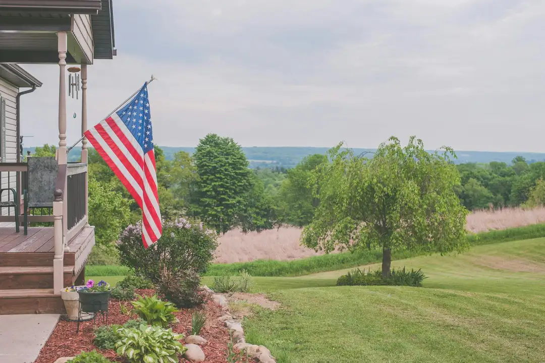 A front porch with an American flag hanging beside a well-kept yard, trees, and a distant open field on a sunny day.