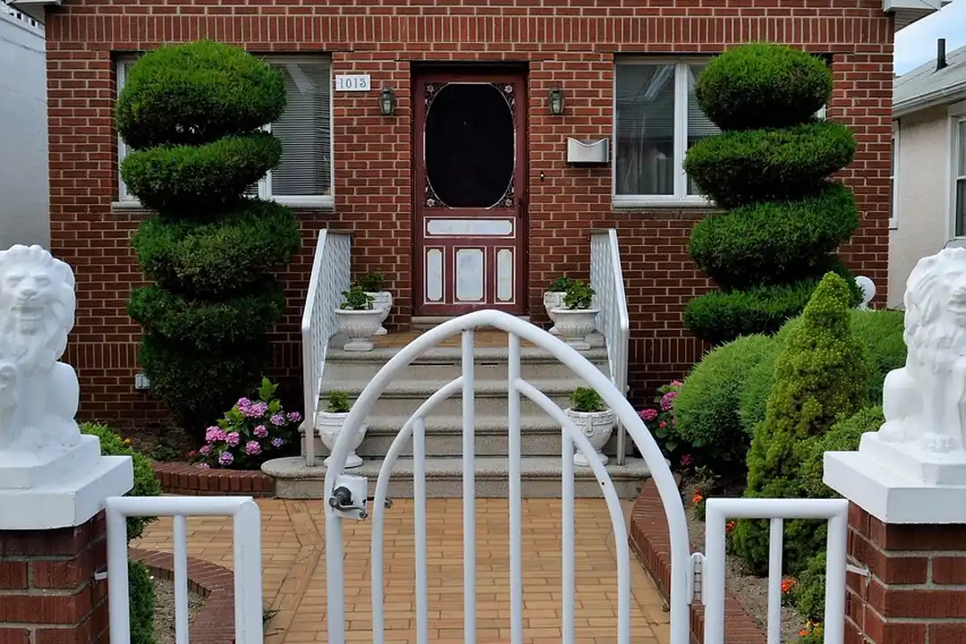 Brick house with manicured evergreen topiaries, a white curved gate, and decorative white bust statues flanking the front entrance.