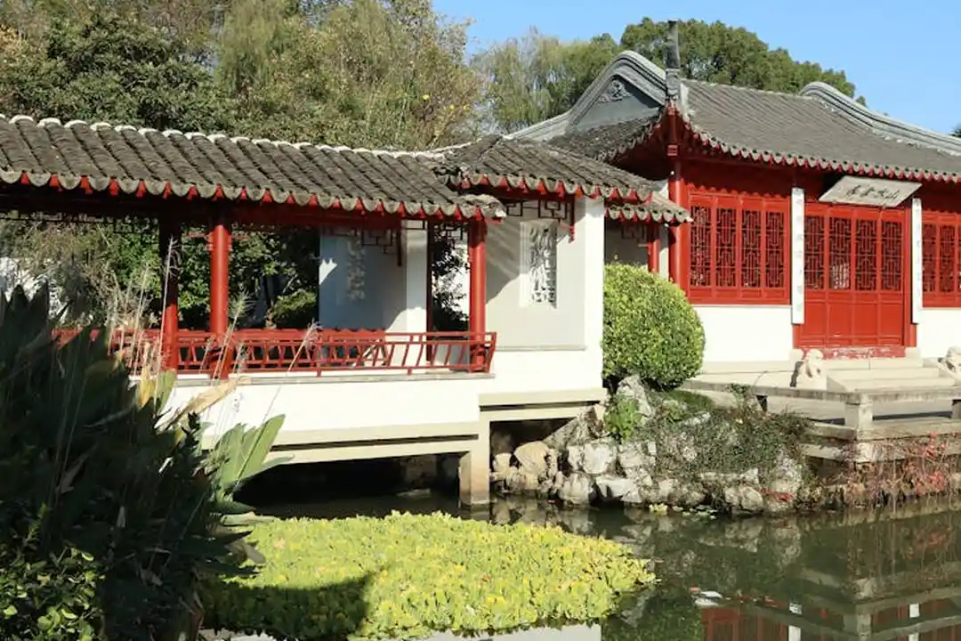 A traditional Chinese-style pavilion over a tranquil pond in a garden, with red accents and a tiled roof.