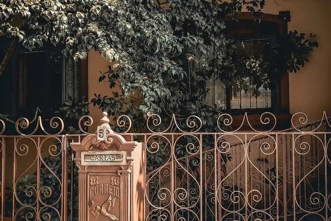 Ornate wrought-iron gate with decorative scrollwork and a small mailbox, set against a leafy background.