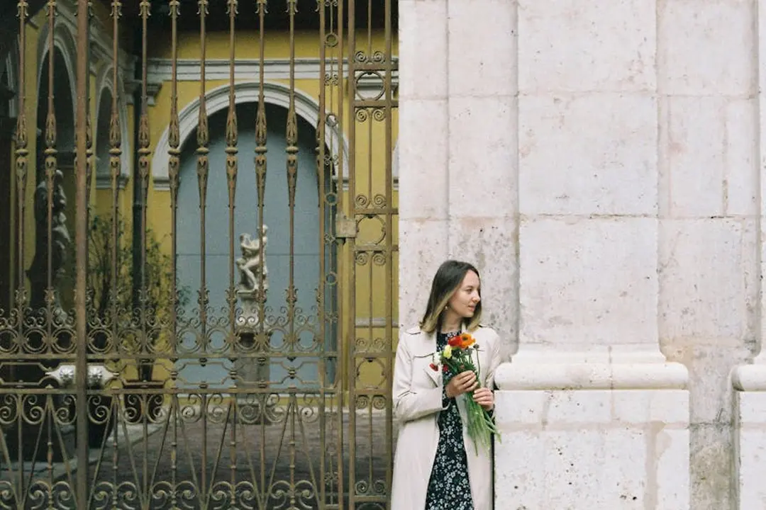 A woman stands beside a decorative wrought-iron gate of a gated community, holding a bouquet of flowers, with a stone column and a yellow building in the background.