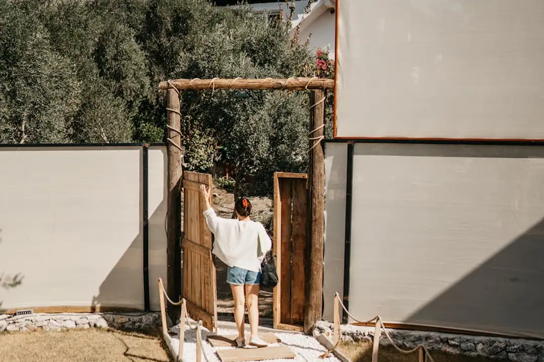 A person enters through an open wooden gate into a gated community, flanked by privacy walls and a gravel path.