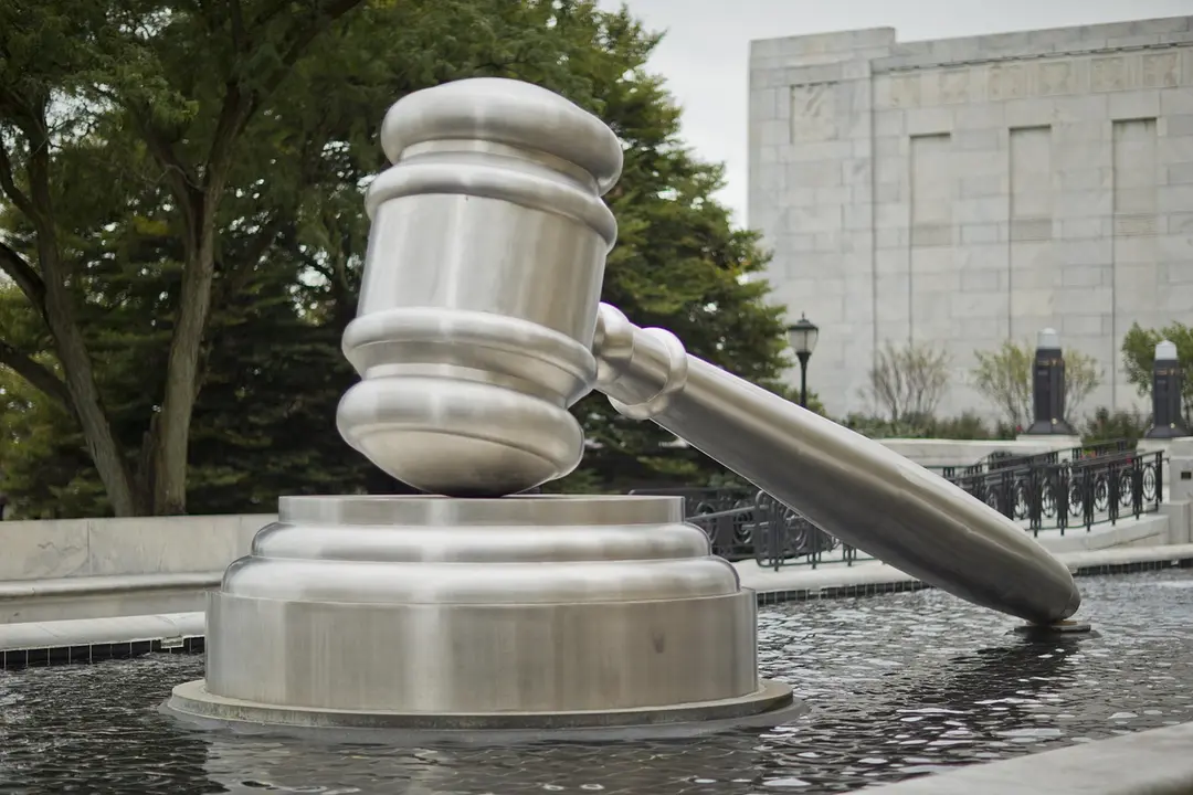 Close-up of a metallic judge's gavel resting on its base above a shallow pool, with a courthouse-style building in the background.