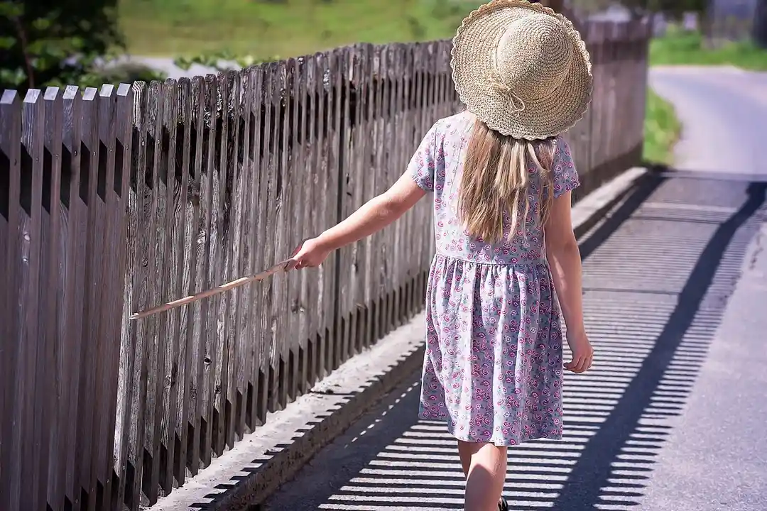 A young girl wearing a sun hat walks along a wooden fence beside a sidewalk on a sunny day.