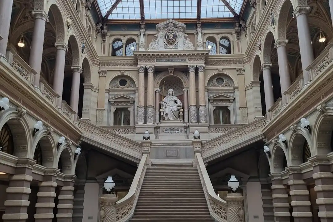 Inside a historic building with a grand marble staircase, arched hallways, columns, and statues, arranged symmetrically under a glass skylight.