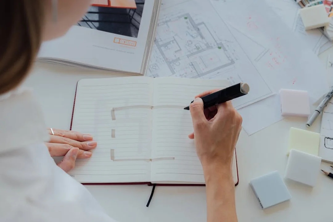 Top-down view of a designer’s hands at a notebook, with architectural plans, color swatches, and draft materials spread across a white desk.