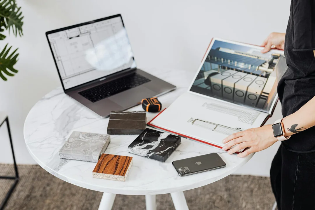 A designer reviews design assets on a laptop and tablet while selecting material swatches on a round white table.