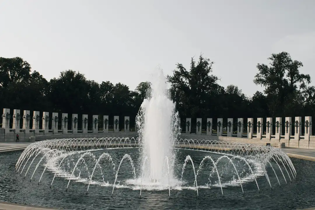 Circular fountain with multiple water jets creating arcs in a stone-bordered plaza, with pillars and trees in the background.