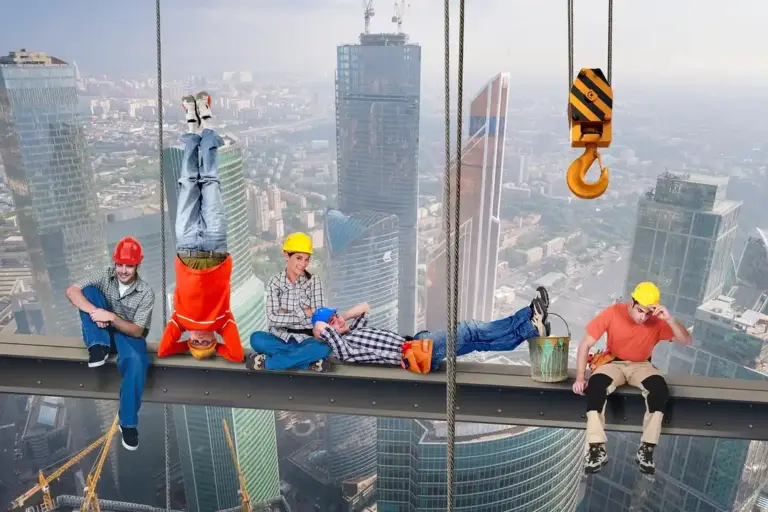 Construction workers wearing hard hats and safety gear sit on a steel beam high above a city skyline during a crane operation.