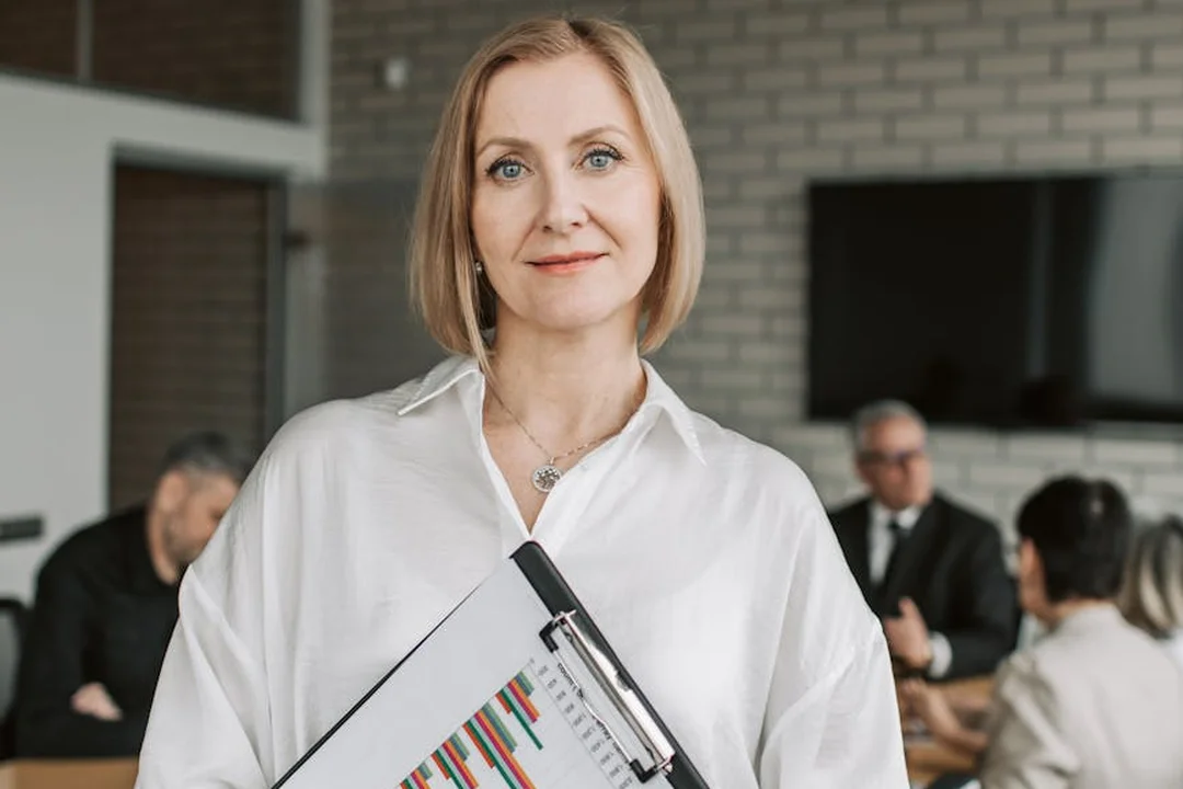 Professional woman holding a folder with documents in a conference room, with colleagues blurred in the background.