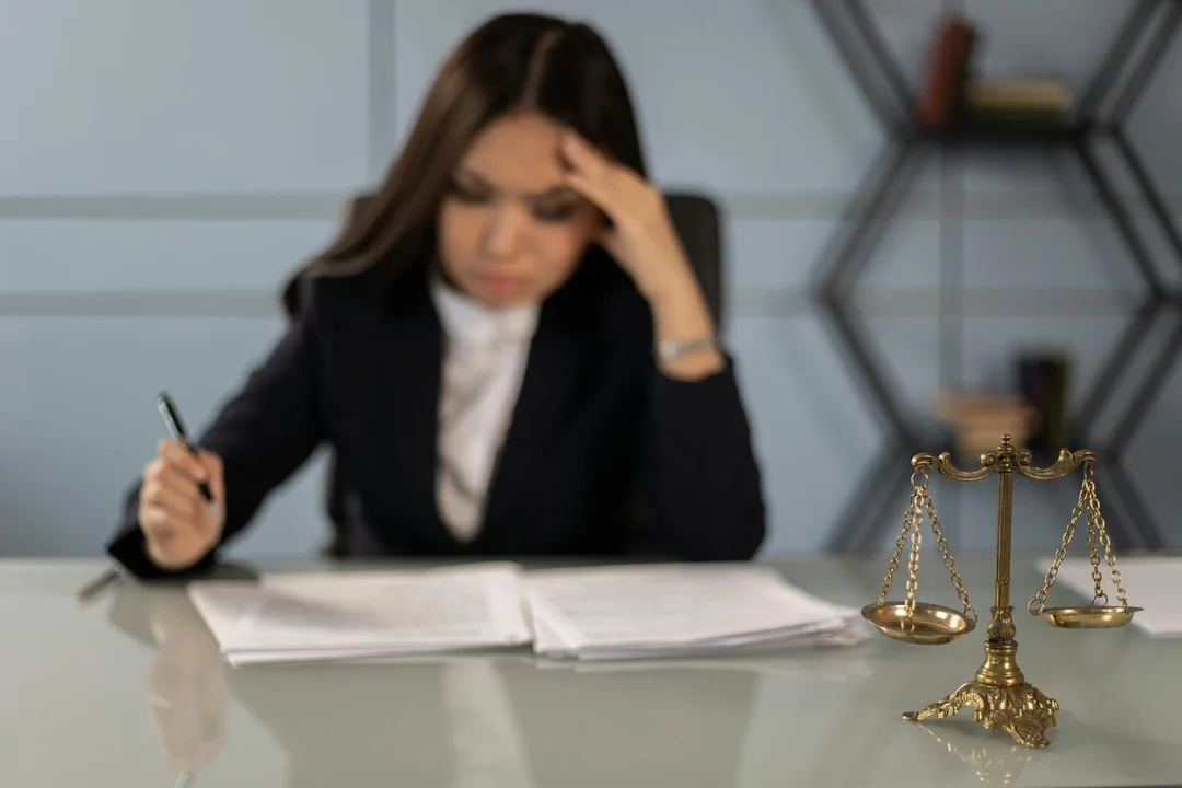 Person in business attire reviews documents at a glass desk with scales of justice nearby.