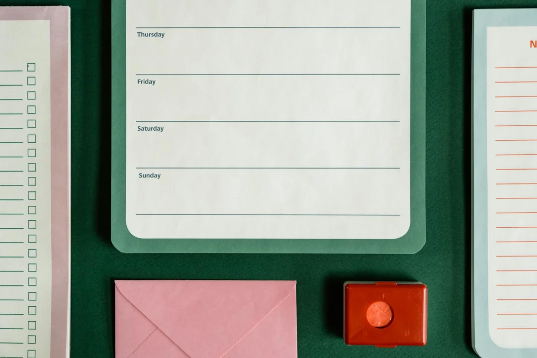 Close-up of a calendar page on a green desk, with a pink envelope and a red stamp pad nearby, and other stationery items.