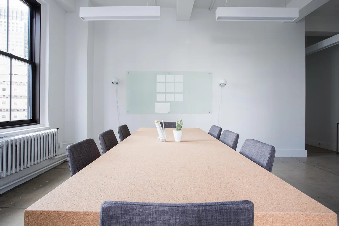 Empty conference room with a long table surrounded by gray chairs, a glass whiteboard on the wall, and large windows to the left.