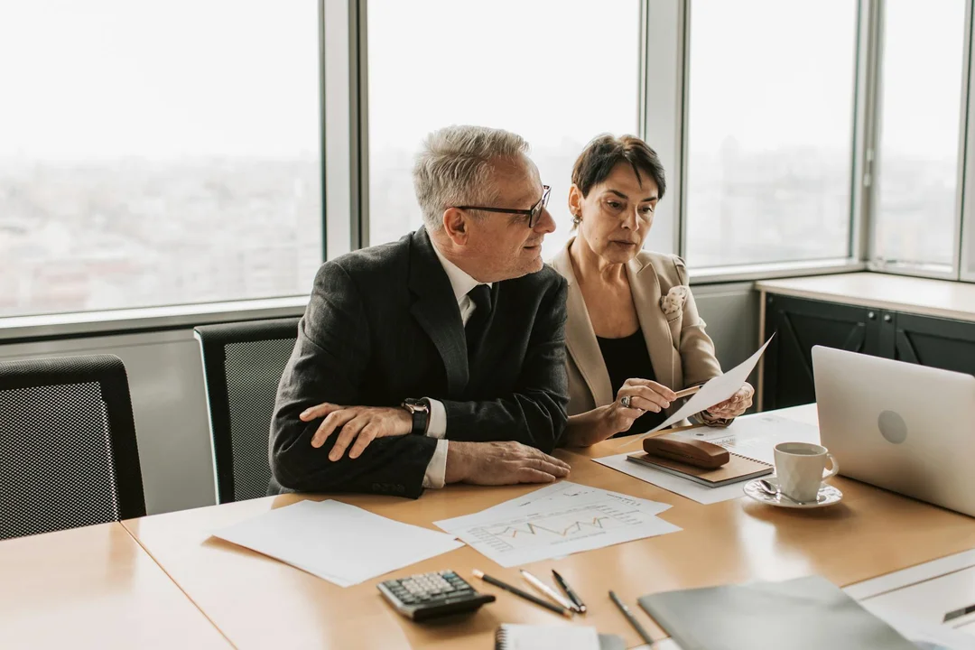 Two professionals sit at a conference table by a large window, reviewing documents and a laptop as they work on an HOA budget.