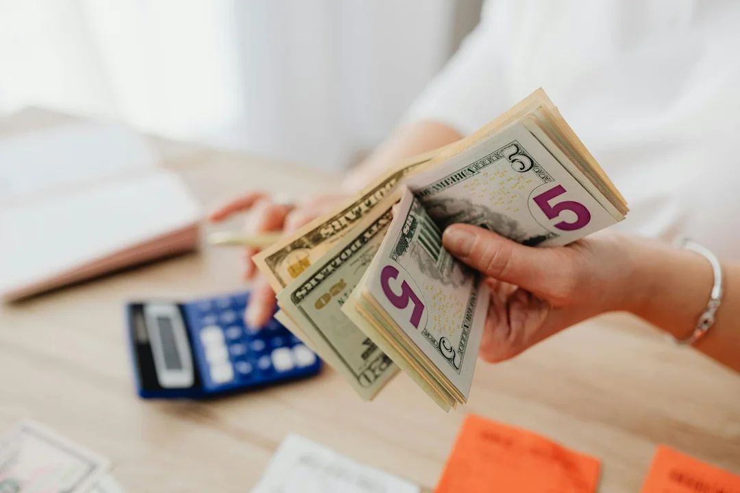 Close-up of hands counting money with a calculator, papers, and sticky notes on a desk