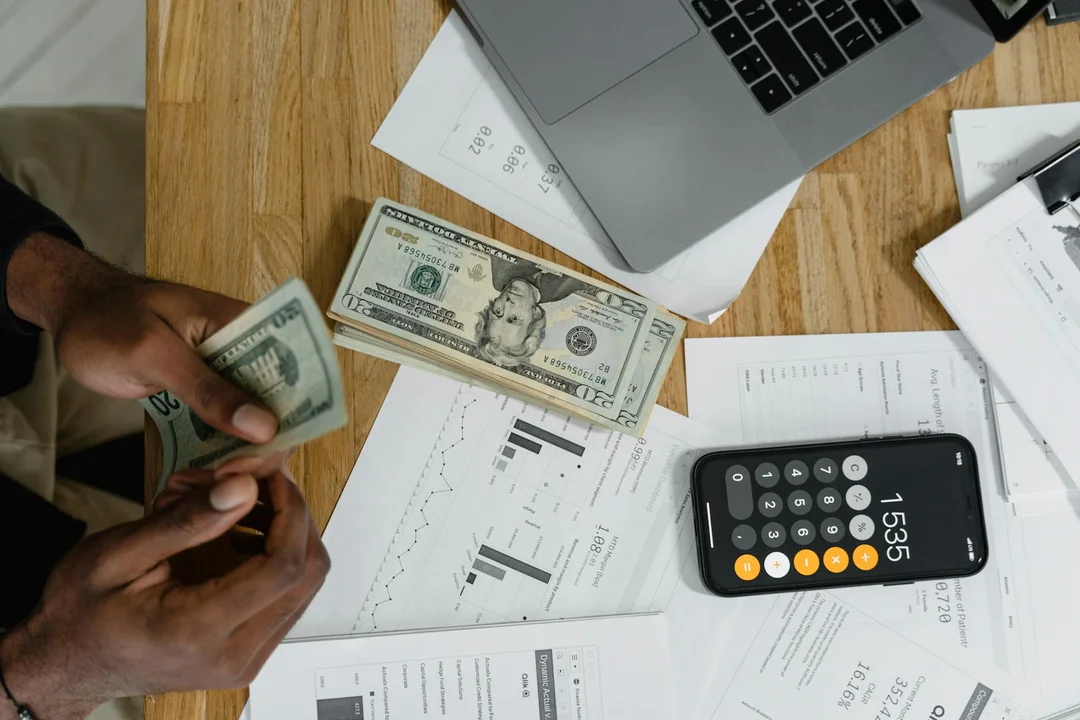 Person counting cash and reviewing financial documents with a calculator and laptop on a wooden desk.