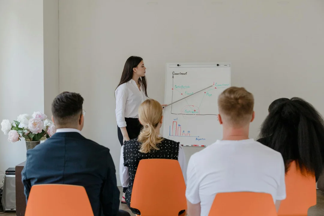 A woman presenter stands at a whiteboard presenting to a small audience seated in orange chairs in a conference room.