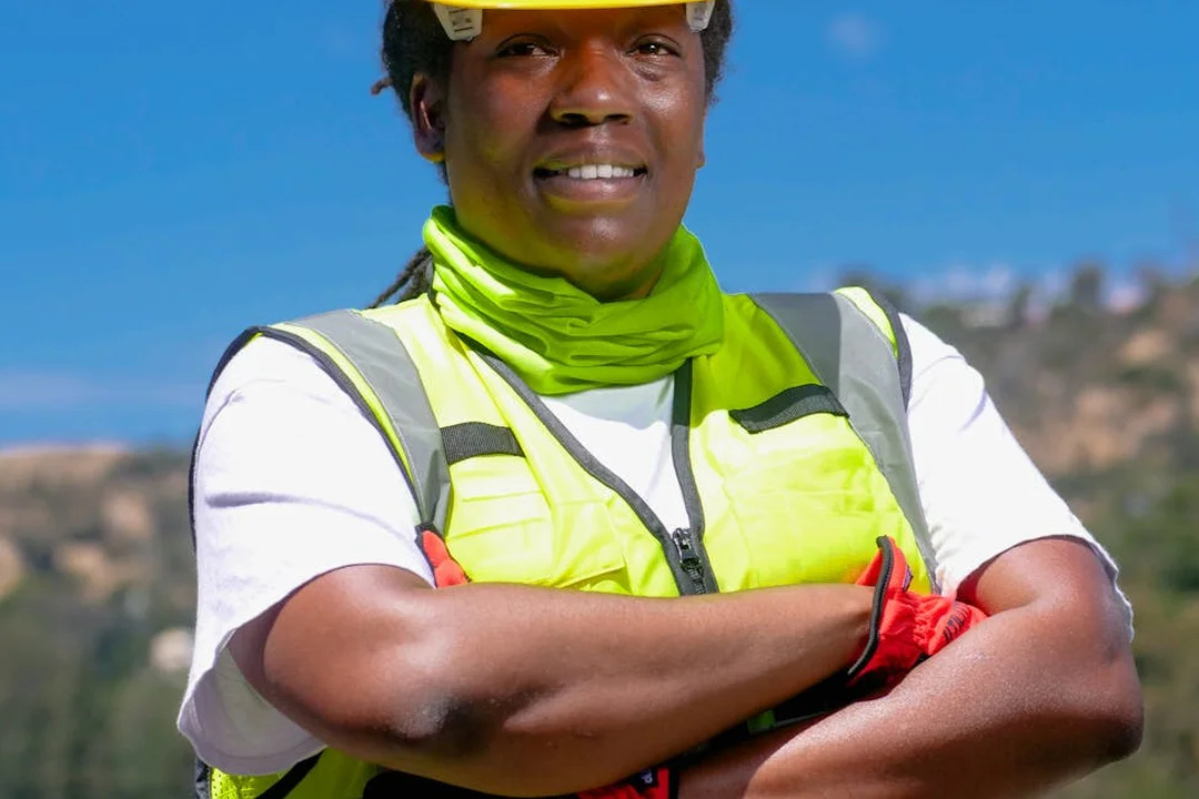 Construction worker wearing a neon safety vest and helmet, arms crossed outdoors with a blue sky