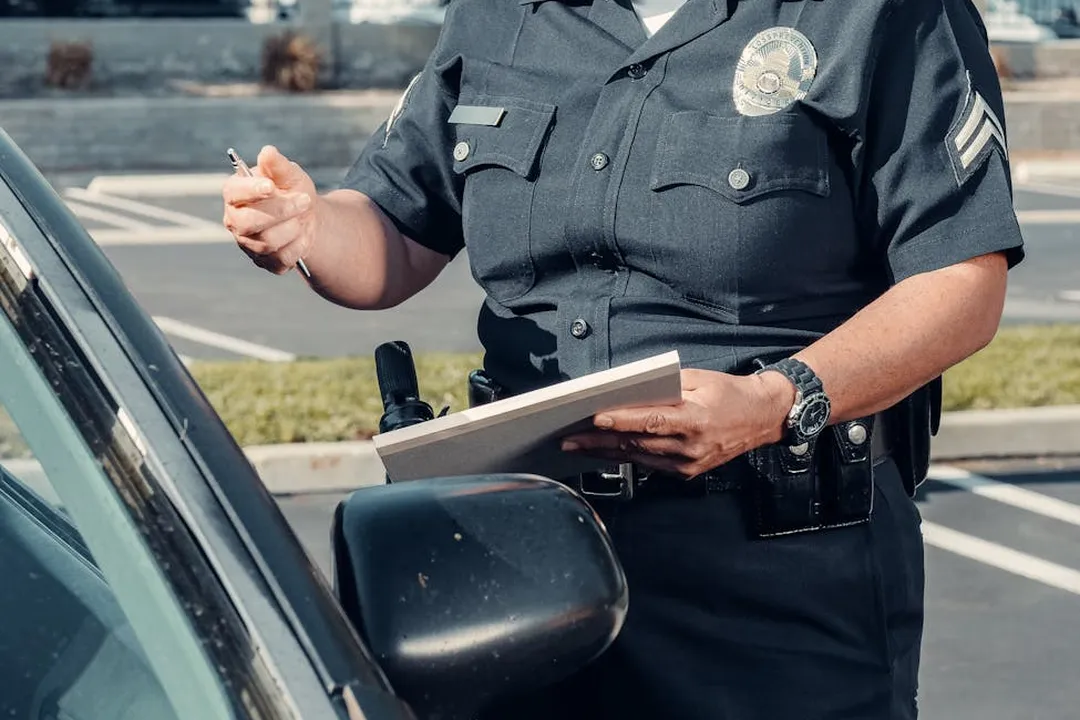 A uniformed officer writes notes at a car window, holding a clipboard and pen.