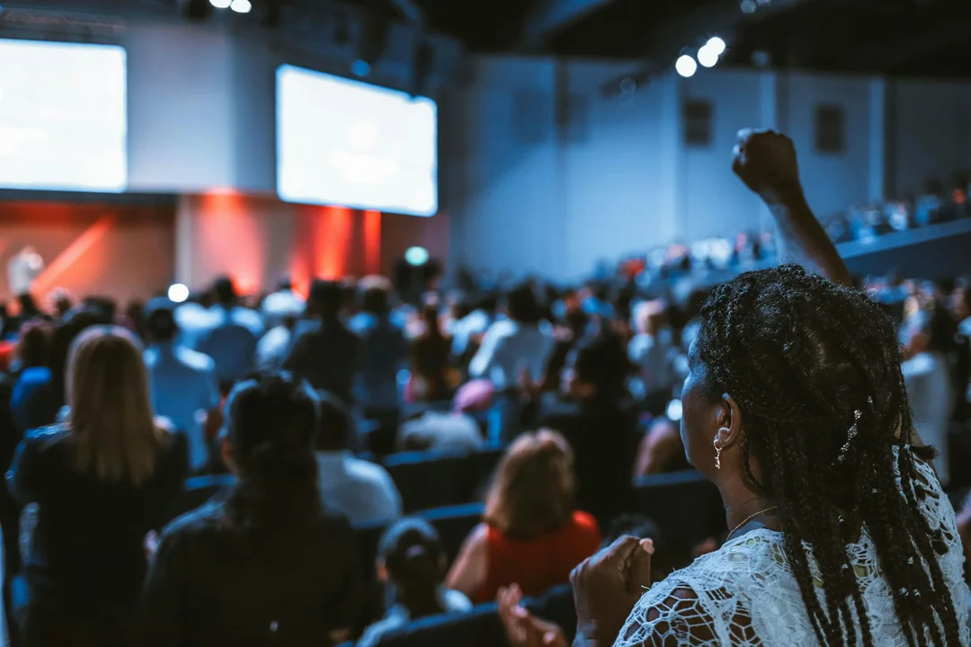 Audience at a conference or HOA meeting, with attendees seated in a large room and a person in the foreground raising a fist; stage screens and lighting visible in the background.