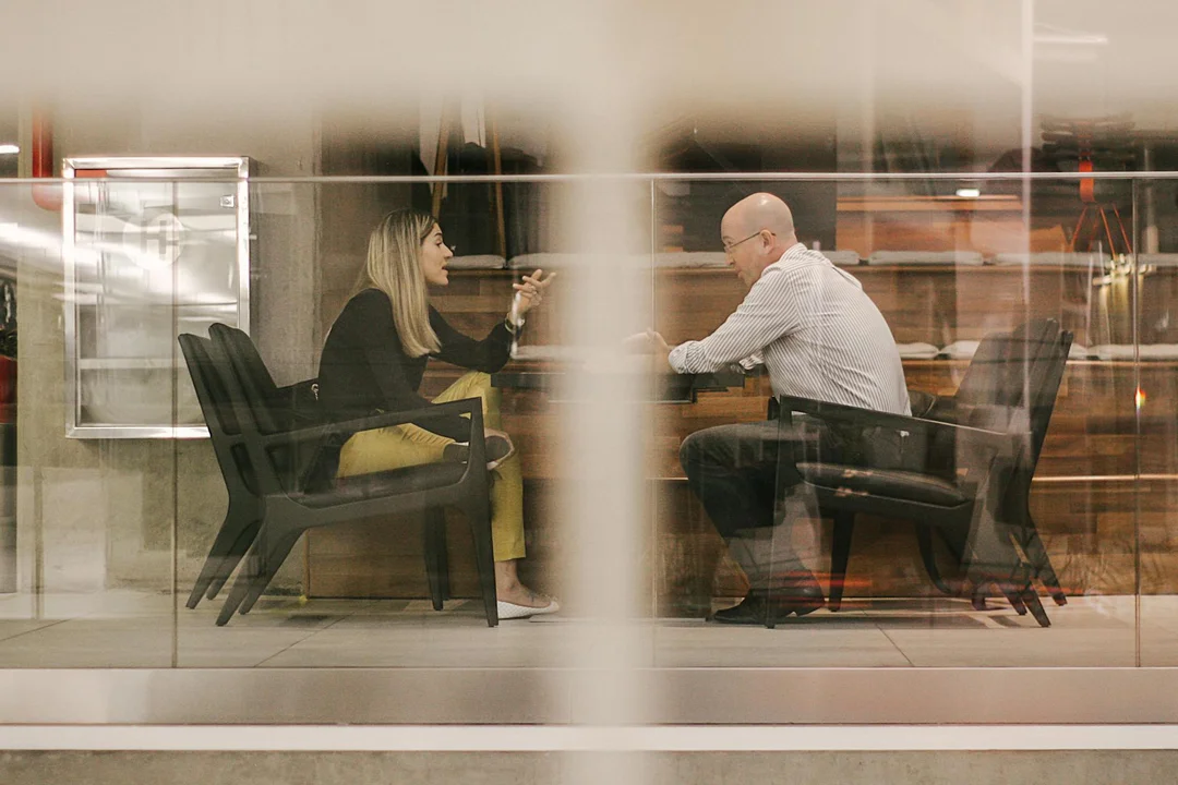 Two homeowners sit across from each other in a glass-walled conference room, discussing strategies to rally neighbors against neglect of common areas.