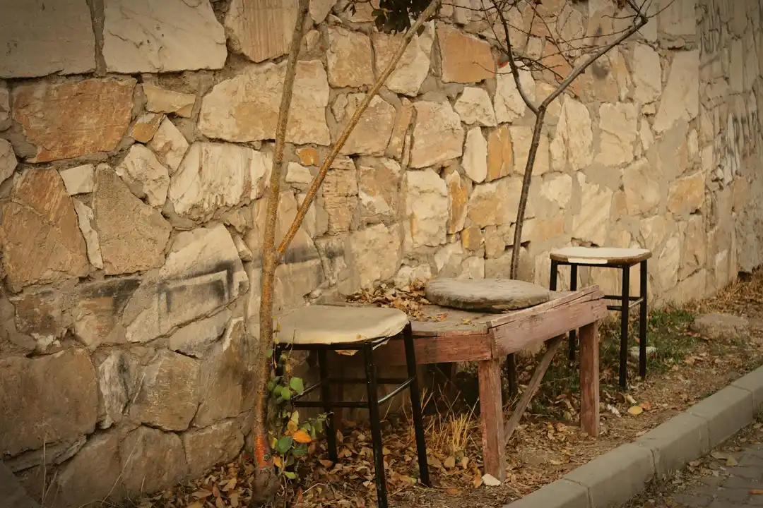 Outdoor seating area along a stone wall with a wooden table and metal stools, beside a sidewalk, with fallen leaves on the ground.
