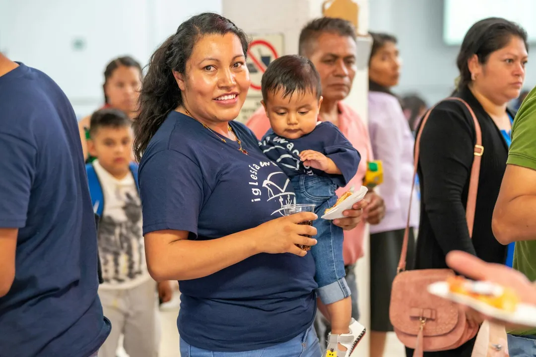 A smiling woman in a blue shirt holds a child while standing among others in a busy HOA community center, with people waiting in line and plates of food visible.