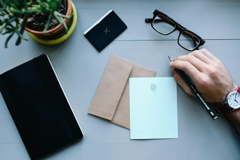 A person writes with a black pen at a tidy desk, with envelopes, a small notebook, a potted plant, a tablet, and a pair of glasses nearby.